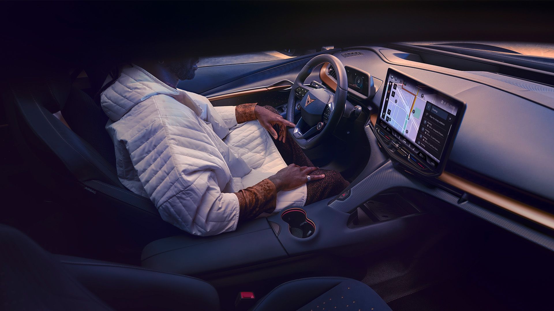 Moderný interiér automobilu s dotykovou obrazovkou na pravej strane vedľa volantu v palubnej doske. Vodič v bielej prešívanej bunde sa dotýka operadla sedadla.