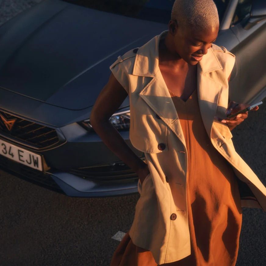 Pohľad na ženu s blond vlasmi, ktorá stojí vedľa tmavosivého automobilu a drží mobilný telefón. Žena je oblečená v svetlom plášti a šatách.