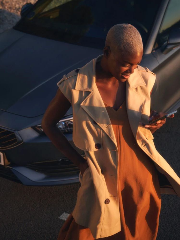 Pohľad na ženu s blond vlasmi, ktorá stojí vedľa tmavosivého automobilu a drží mobilný telefón. Žena je oblečená v svetlom plášti a šatách.