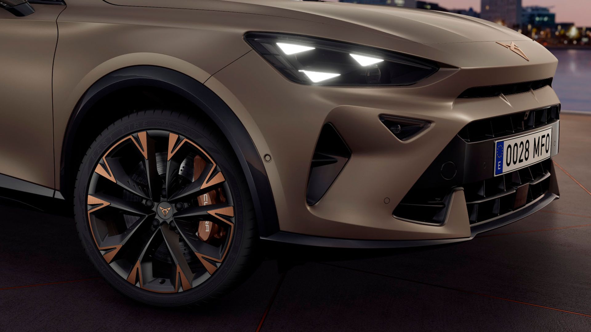 Detailný záber na prednú časť moderného vozidla značky CUPRA so zapnutými prednými svetlometmi. Auto má elegantný matný béžový lak a čierne disky s bronzovými detailmi. V pozadí je rozostrená panoráma meste..