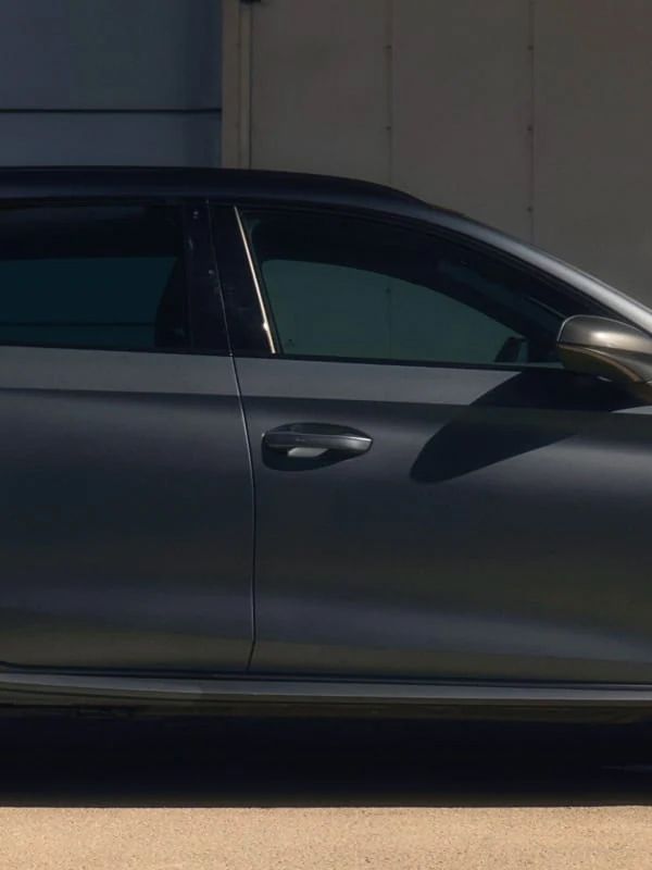 Muž stojí vedľa sivého CUPRA Leon Sportstourer zaparkovaný pred modernou budovou, drží v ruke tablet a má oblečený športový outfit. Pozadie je minimalistické s niekoľkými kovovými prvkami.