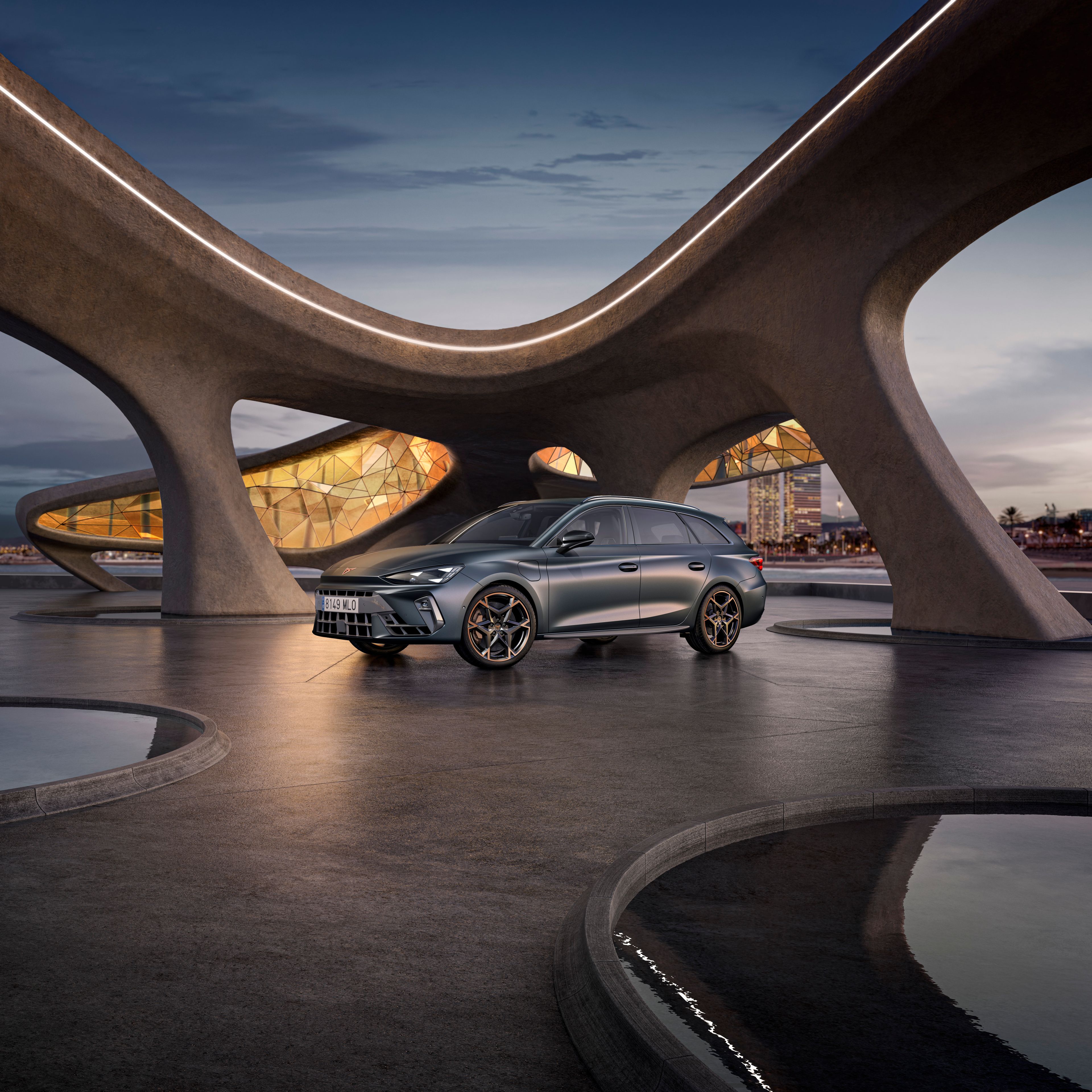 CUPRA Leon Sportstourer zaparkovaný pri modernej sklenenej budove pri mori, v pozadí vidieť pláž a mesto.