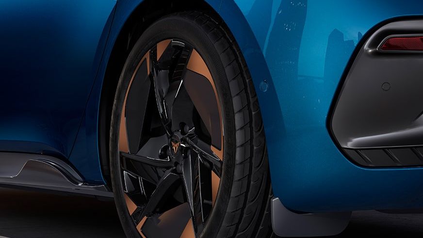 Časť zadných partií modrého automobilu zameraná na moderné koleso. Na pozadí sa odrážajú obrysy budov. Koleso má výrazný dizajn s geometrickými tvarmi.