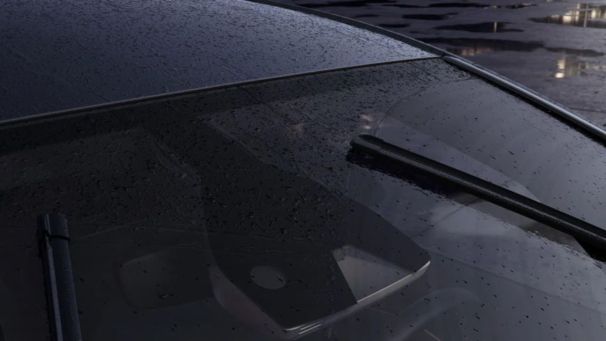 Záber na predné sklo auta, na ktorom sú kvapky dažďa a zapnuté stierače. V pozadí vidieť rozmazané okolité prostredie, pravdepodobne parkovisko.