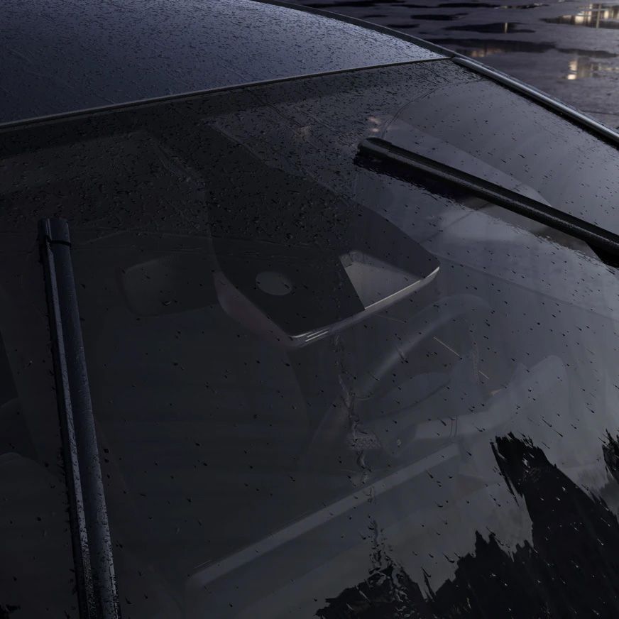 Záber na predné sklo auta, na ktorom sú kvapky dažďa a zapnuté stierače. V pozadí vidieť rozmazané okolité prostredie, pravdepodobne parkovisko.