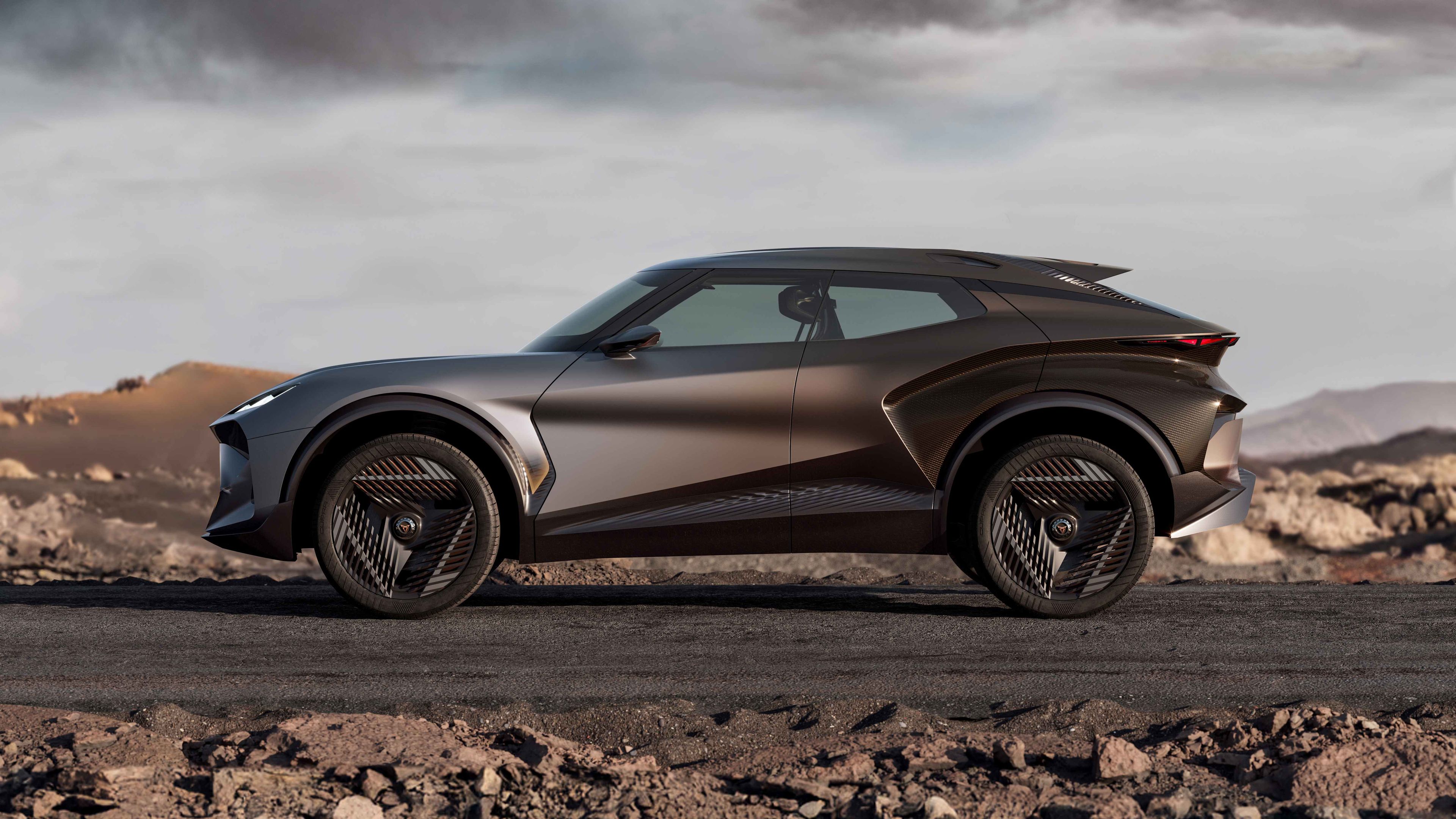 Luxusné, futuristické SUV v kovovo šedej farbe stojí na pustej ceste so skalnatým pohorím v pozadí. Auto má moderný dizajn s úzkymi, šikmými svetlami a veľkými kolesami. Obloha je zatiahnutá s dramatickou atmosférou.