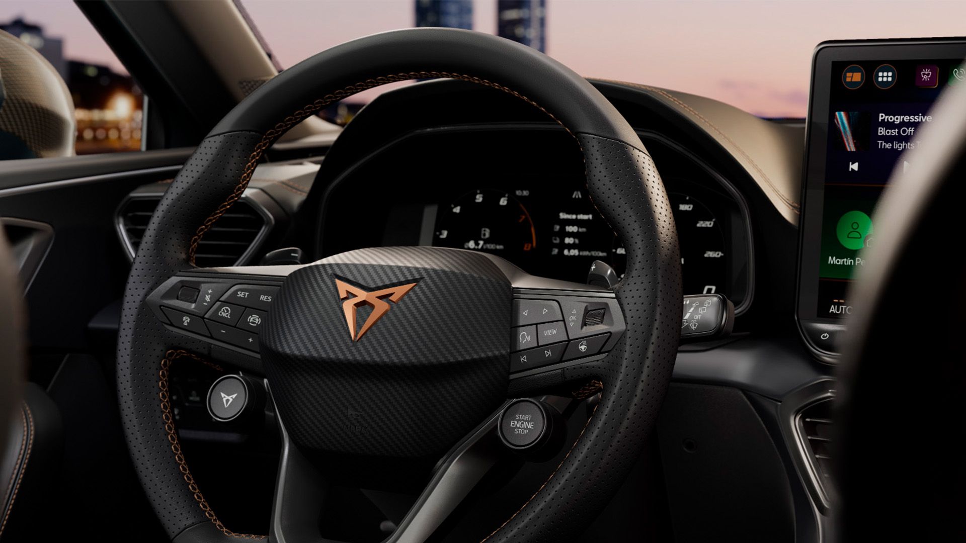 Detailný na multifunkčný volant značky CUPRA.Za volantom je digitálny displej prístrojového panela. Volant je potiahnutý čiernou kožou s oranžovým prešívaním. V pozadí je nejasné mestské prostredie pri západe slnka.