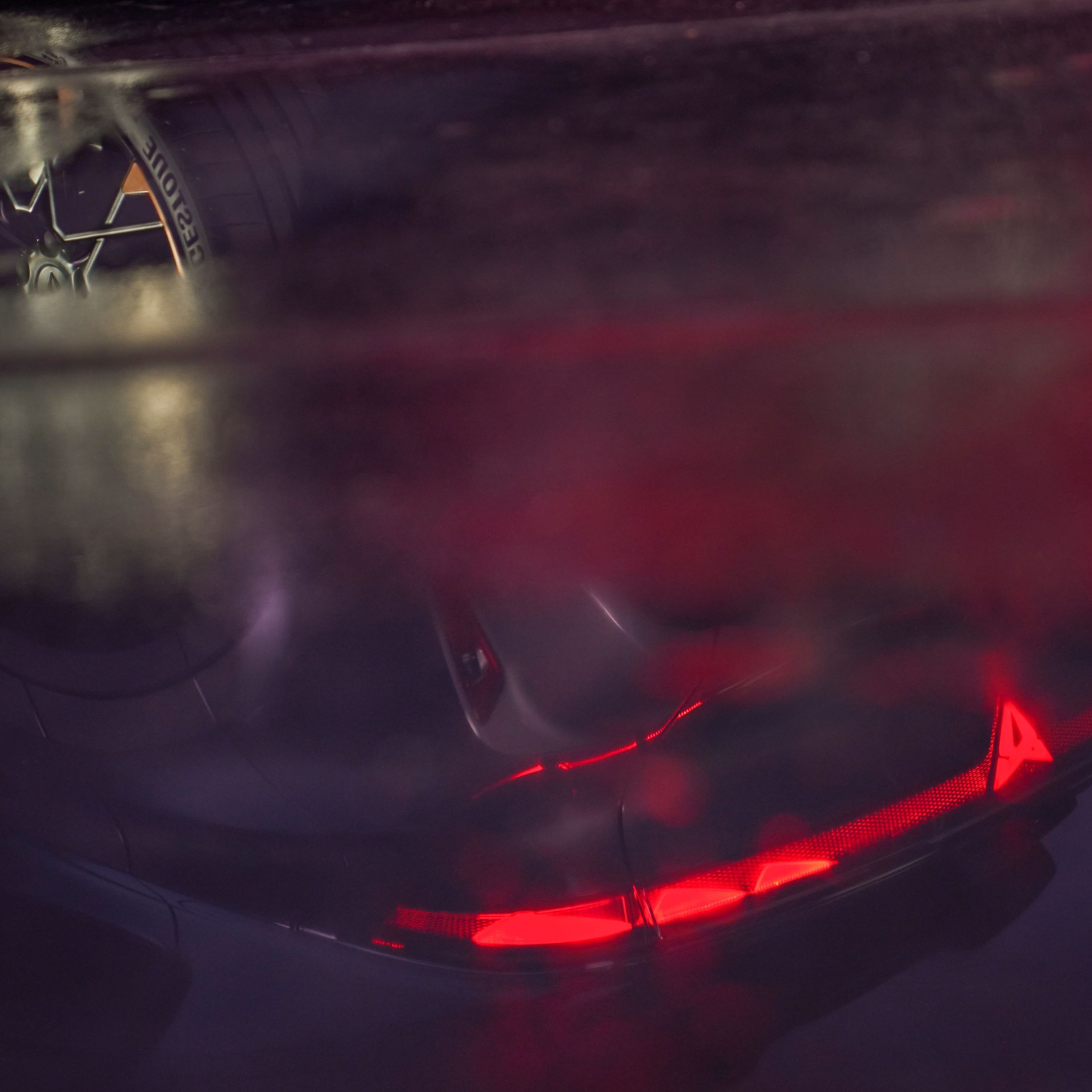 Pohľad na tmavú, mokrú podlahu, v ktorej sa odráža nový model Raval. Detail na zasvietené zadné svetlá.