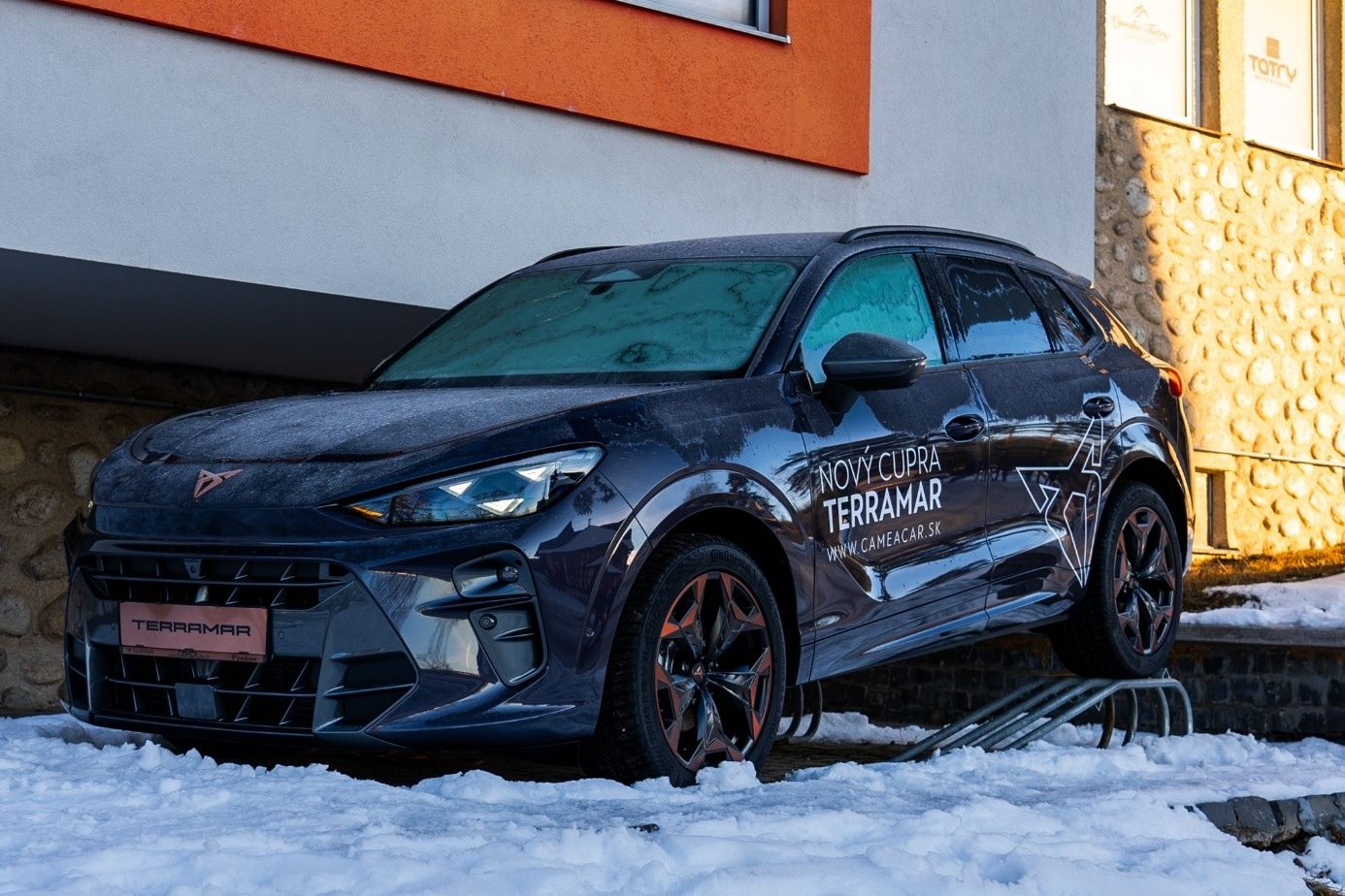 CUPRA Terramar je zaparkované na zasneženej ceste pred budovou a je pokryté ľahkou vrstvou snehu. Má tmavomodrú farbu a čierne disky kolies s bronzovými detailmi. Na dverách sú nápisy "Nový Cupra Terramar".