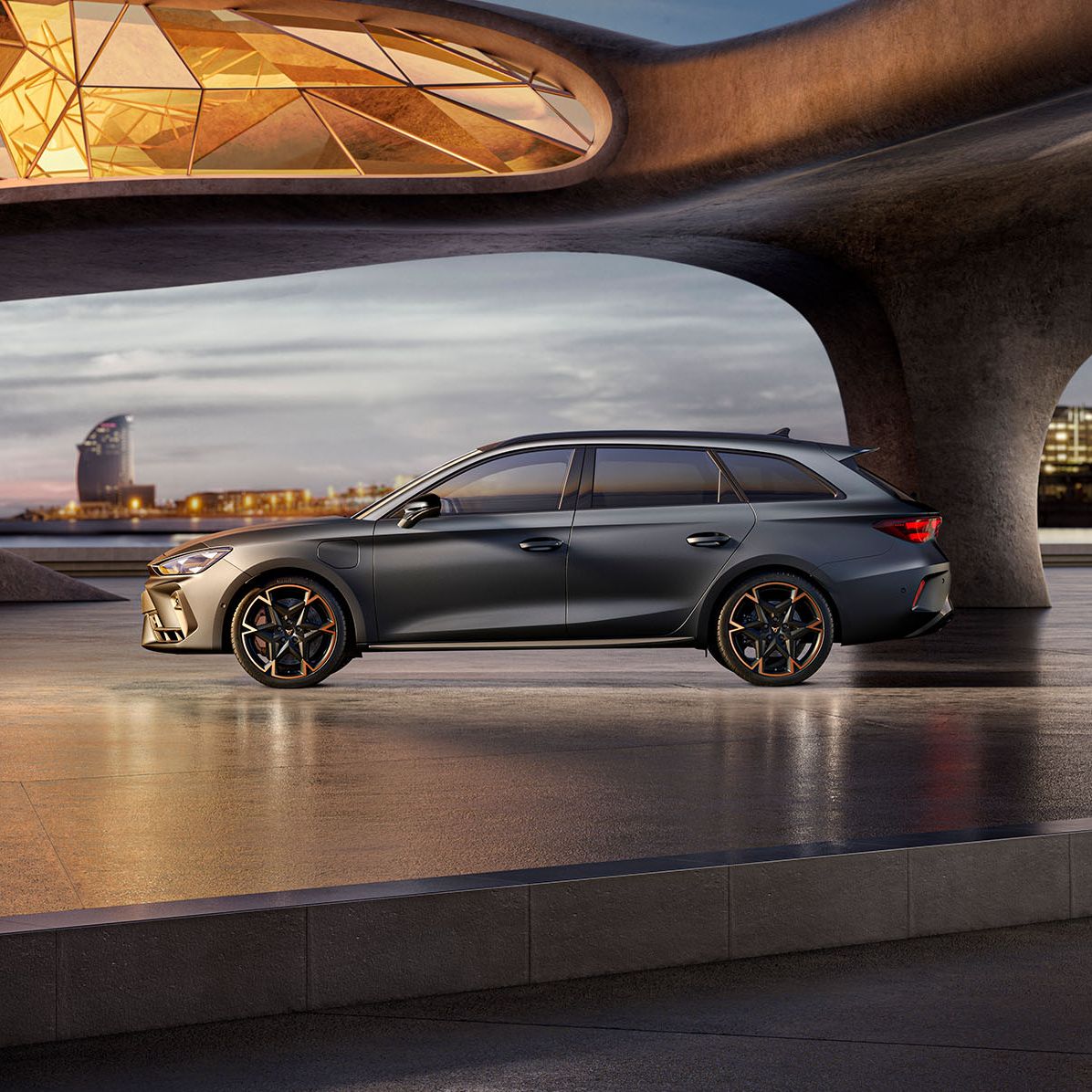 CUPRA Leon Sportstourer stojí na modernom námestí pri umeleckej architektúre. Priestor osvetľuje sklenená strecha, ktorá vytvára zaujímavé svetelné efekty. V pozadí sú budovy a mestská scenéria.