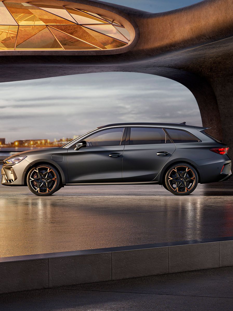 CUPRA Leon Sportstourer stojí na modernom námestí pri umeleckej architektúre. Priestor osvetľuje sklenená strecha, ktorá vytvára zaujímavé svetelné efekty. V pozadí sú budovy a mestská scenéria.