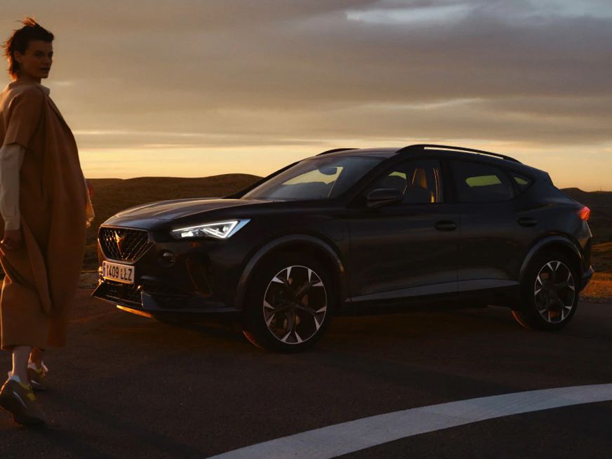 Žena stojí vedľa zaparkovaného športového auta na ceste počas západu slnka. Krajina v pozadí je osvetlená teplými farbami.