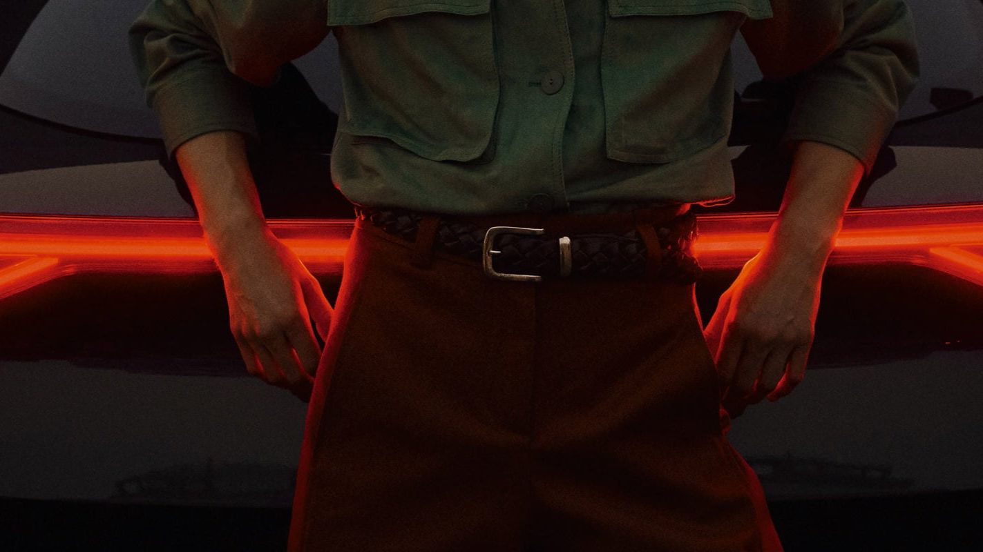 Pohľad zozadu na čierne auto so zapnutými výraznými červenými zadnými svetlami. Pred autom stojí muž v zelenej košeli a hnedých nohaviciach s rukami opretými o vozidlo. 