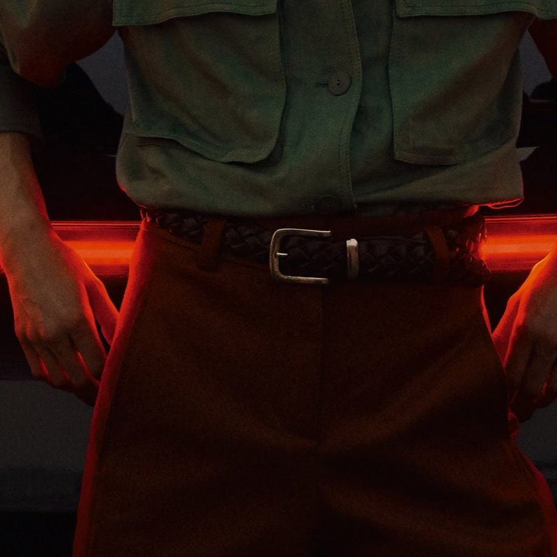 Pohľad zozadu na čierne auto so zapnutými výraznými červenými zadnými svetlami. Pred autom stojí muž v zelenej košeli a hnedých nohaviciach s rukami opretými o vozidlo. 