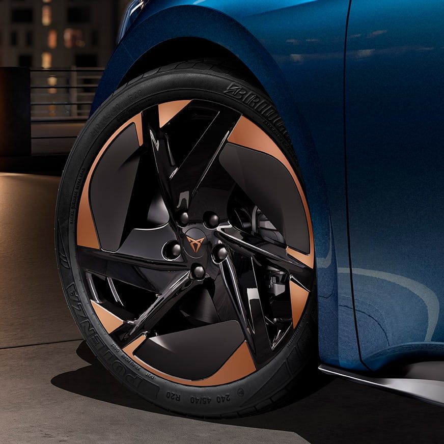 Detailný záber na koleso modrého auta so štýlovými čiernymi diskami a medenými prvkami. V pozadí sú rozmazané mestské budovy.