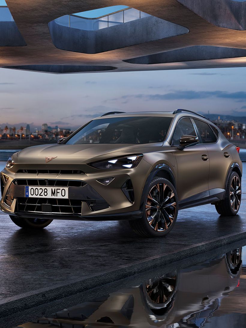 Moderné SUV sa nachádza pod futuristickým, architektonicky zaujímavým mostom vedľa stĺpov za súmraku. V pozadí je panoráma mesta. Auto je zaparkované na mokrej dlažbe, v ktorej sa odráža jeho obraz.