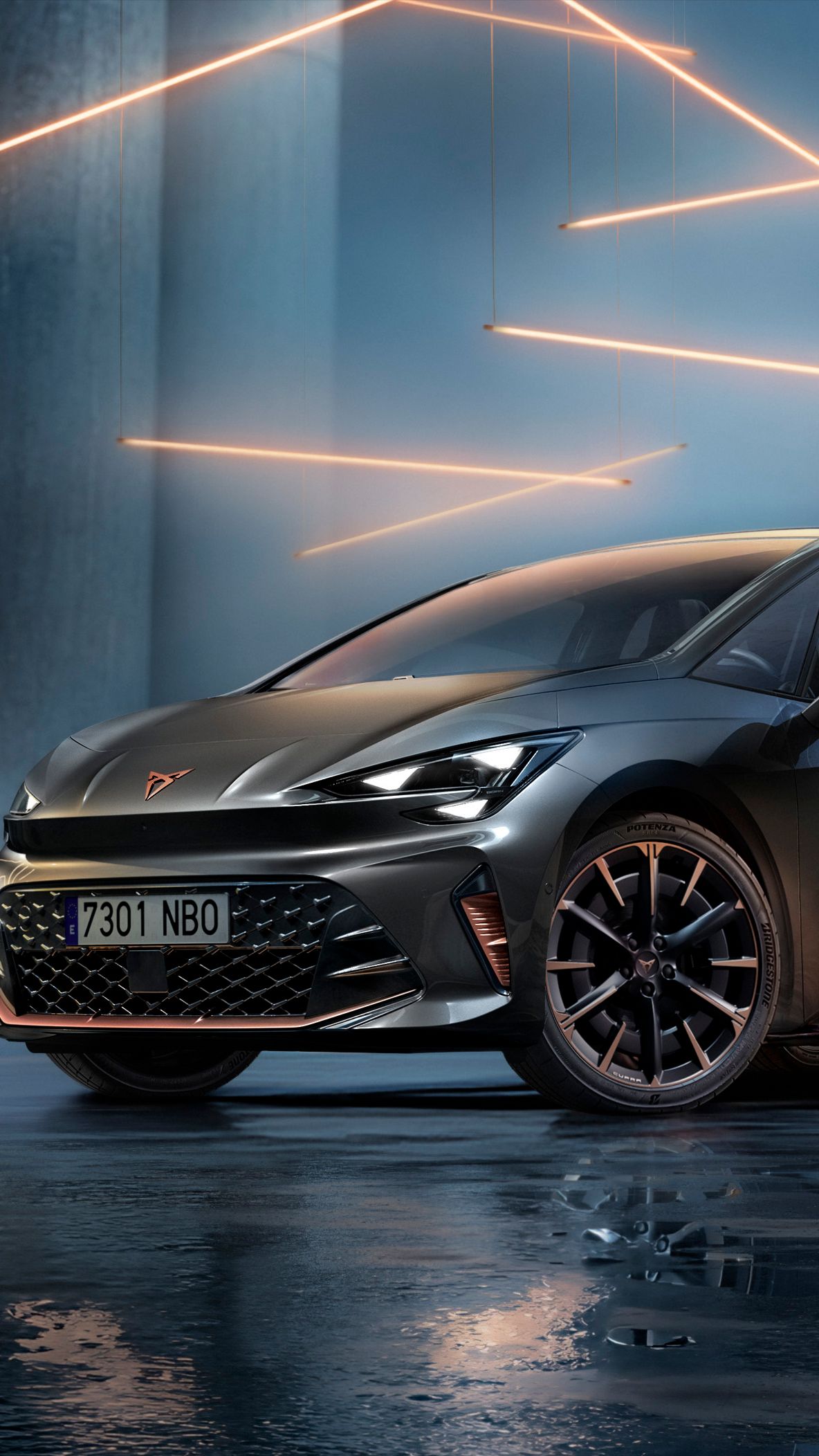 Sivé vozidlo CUPRA Born stojí v modernom, tmavom prostredí s vertikálnymi stĺpmi a svietiacimi LED pásmi. Podlaha je lesklá a odráža vozidlo aj osvetlenie. Auto pôsobí mohutne a elegantne.