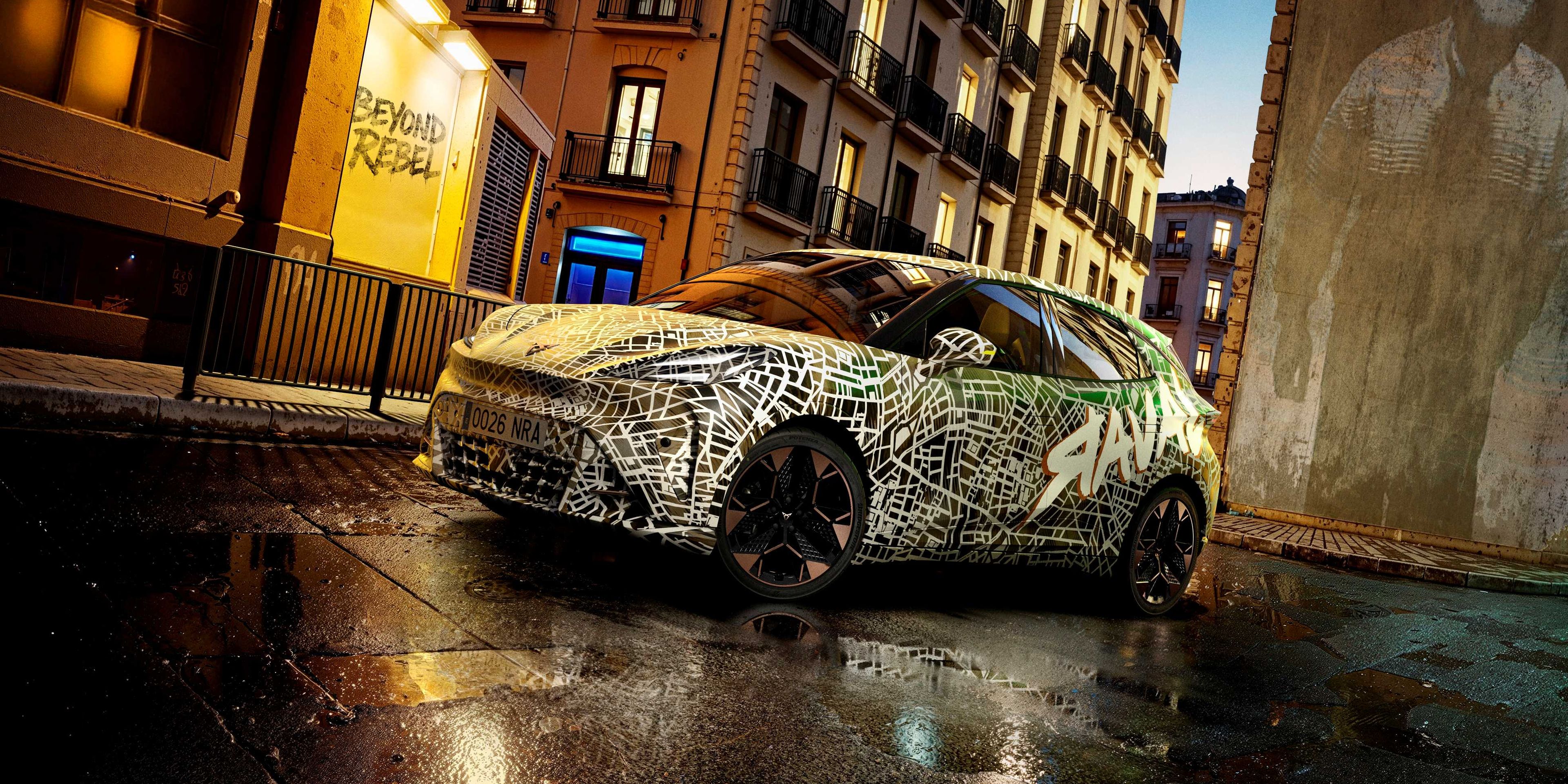 Kamuflovaný model CUPRA stojí na mokrej ceste pred historickou budovou za súmraku.