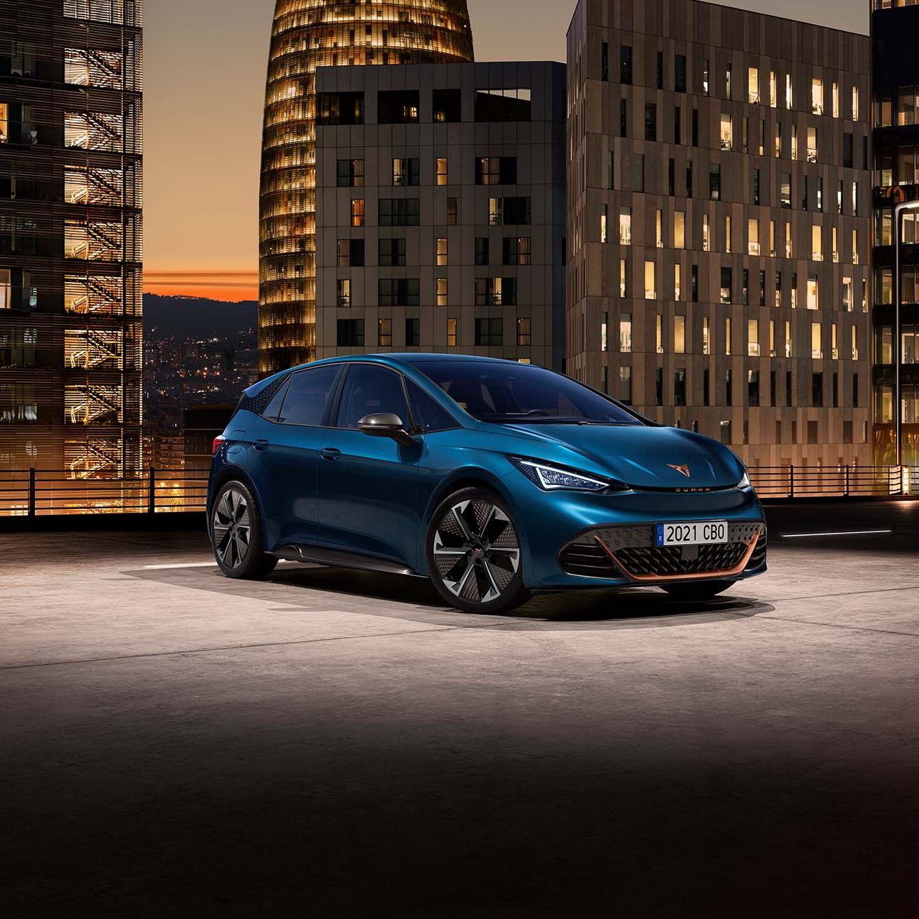 CUPRA Born zaparkovaný na streche parkovacej budovy. V pozadí sú moderné výškové budovy pri západe slnka. Auto je modré.