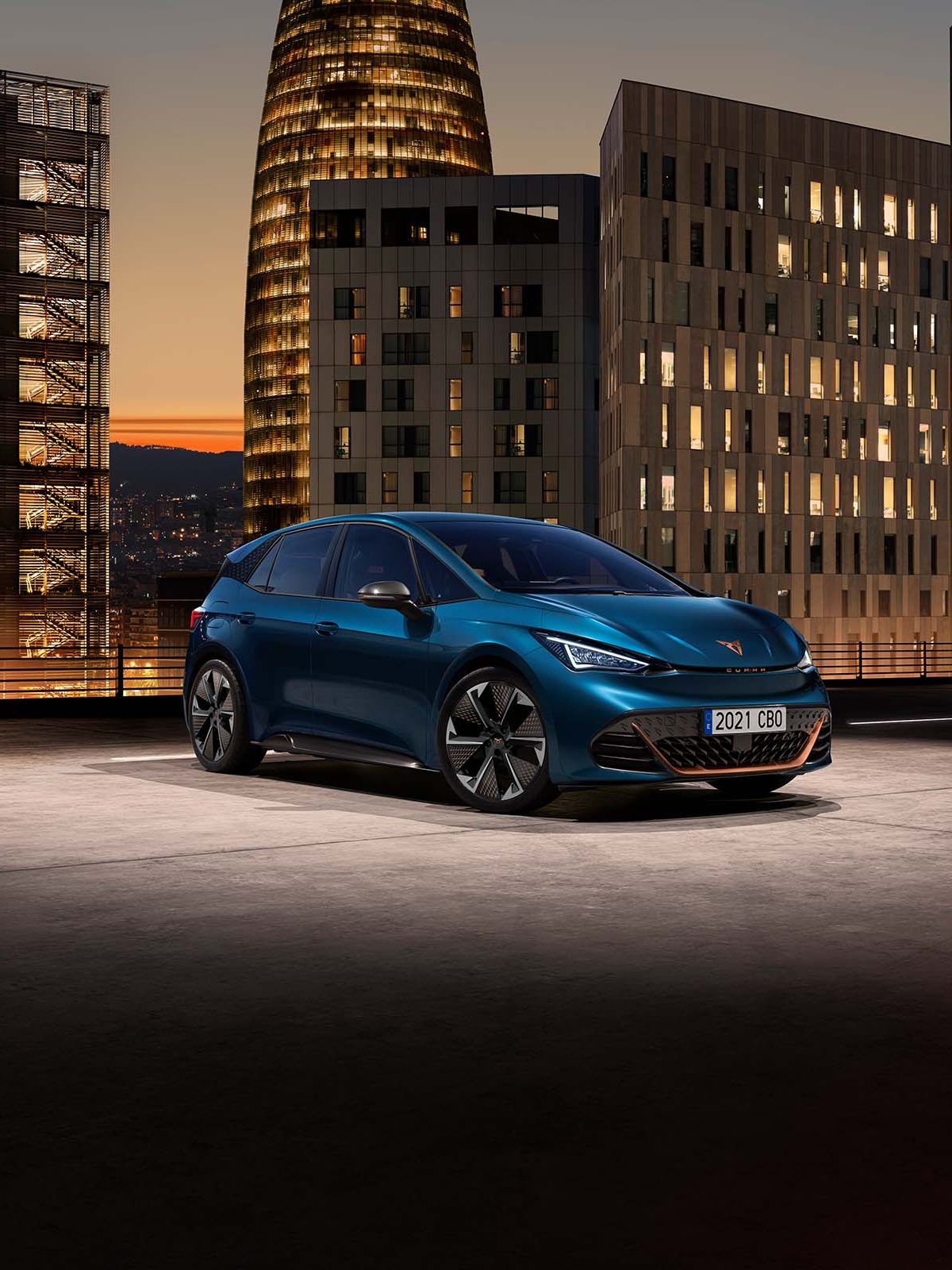 CUPRA Born zaparkovaný na streche parkovacej budovy. V pozadí sú moderné výškové budovy pri západe slnka. Auto je modré.