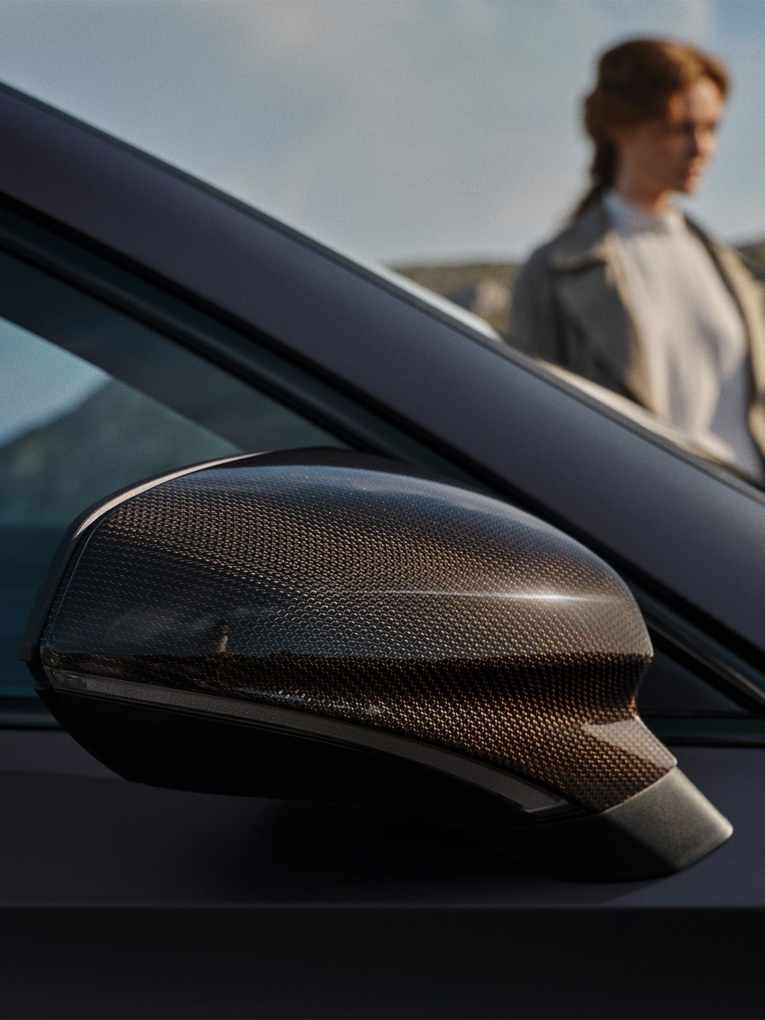 Detail na vonkajšie spätné zrkadlo s krytom z uhlíkovým vlákien. V pozadí je rozmazaná postava ženy stojacej vedľa auta. 