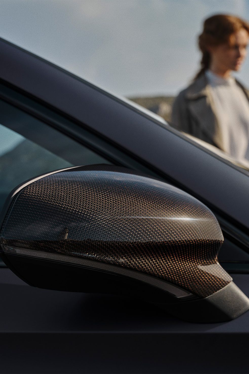 Detail na vonkajšie spätné zrkadlo s krytom z uhlíkovým vlákien. V pozadí je rozmazaná postava ženy stojacej vedľa auta. 