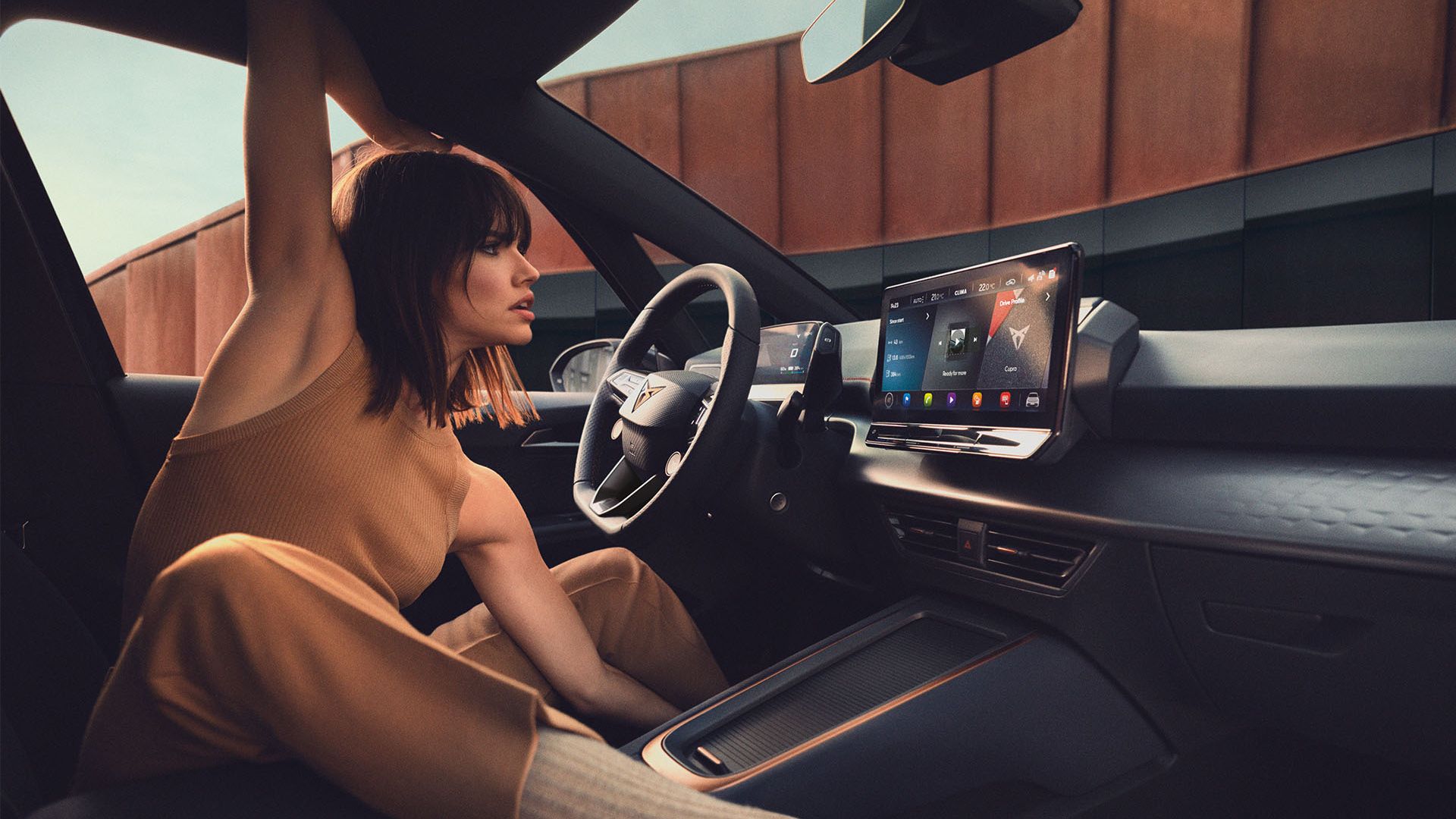 Mladá žena sedí na sedadle vodiča a naťahuje sa. Interiér vozidla je vybavený veľkým dotykovým displejom na palubnej doske.