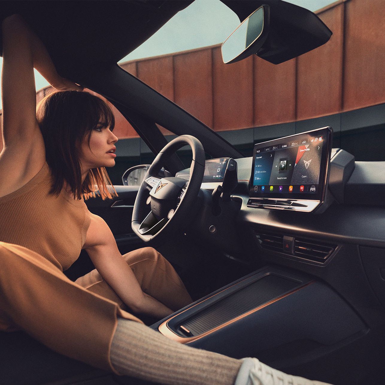 Mladá žena sedí na sedadle vodiča a naťahuje sa. Interiér vozidla je vybavený veľkým dotykovým displejom na palubnej doske.