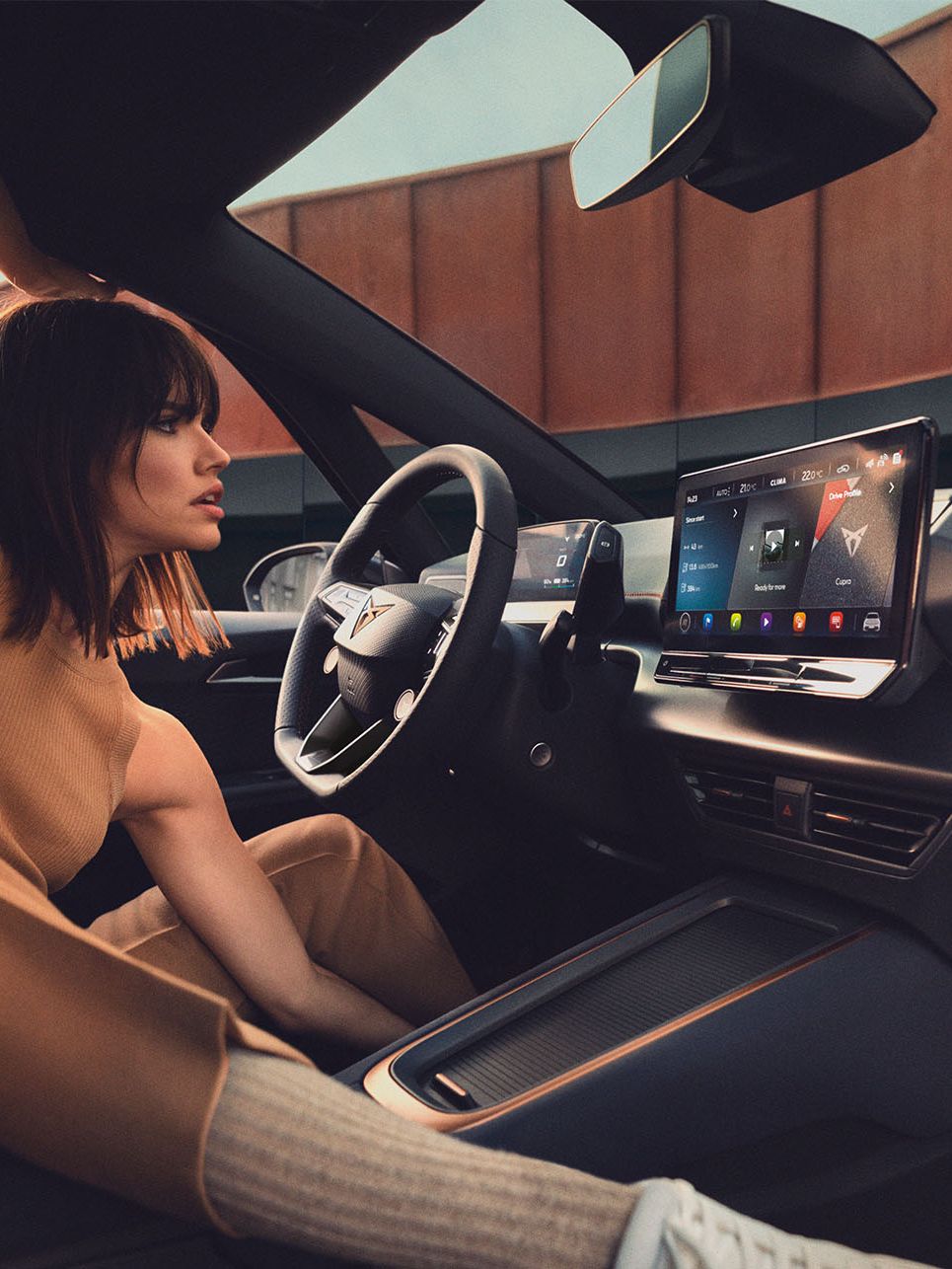 Mladá žena sedí na sedadle vodiča a naťahuje sa. Interiér vozidla je vybavený veľkým dotykovým displejom na palubnej doske.