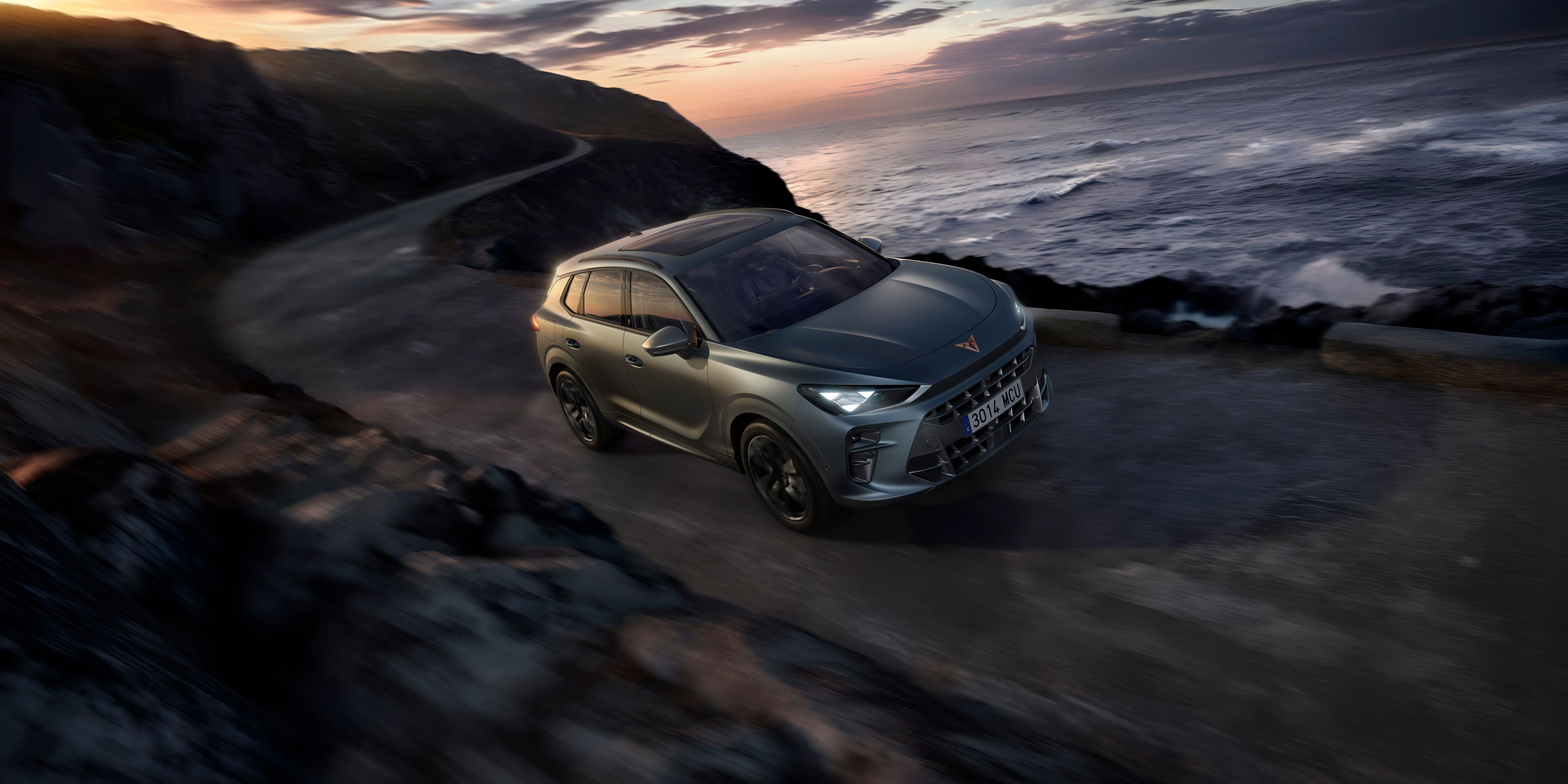 Automobil jazdí po úzkej ceste vedľa pobrežia za súmraku. More je rozbúrené a obloha je zamračená. Autu sa darí aj napriek náročným podmienkam na ceste.