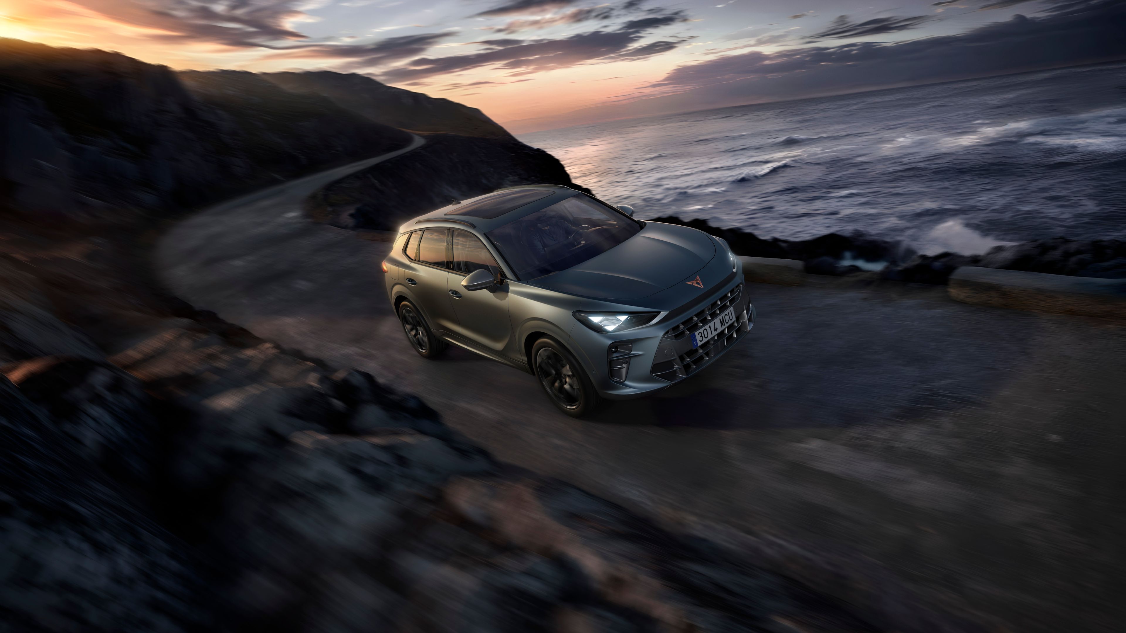 Automobil jazdí po úzkej ceste vedľa pobrežia za súmraku. More je rozbúrené a obloha je zamračená. Autu sa darí aj napriek náročným podmienkam na ceste.