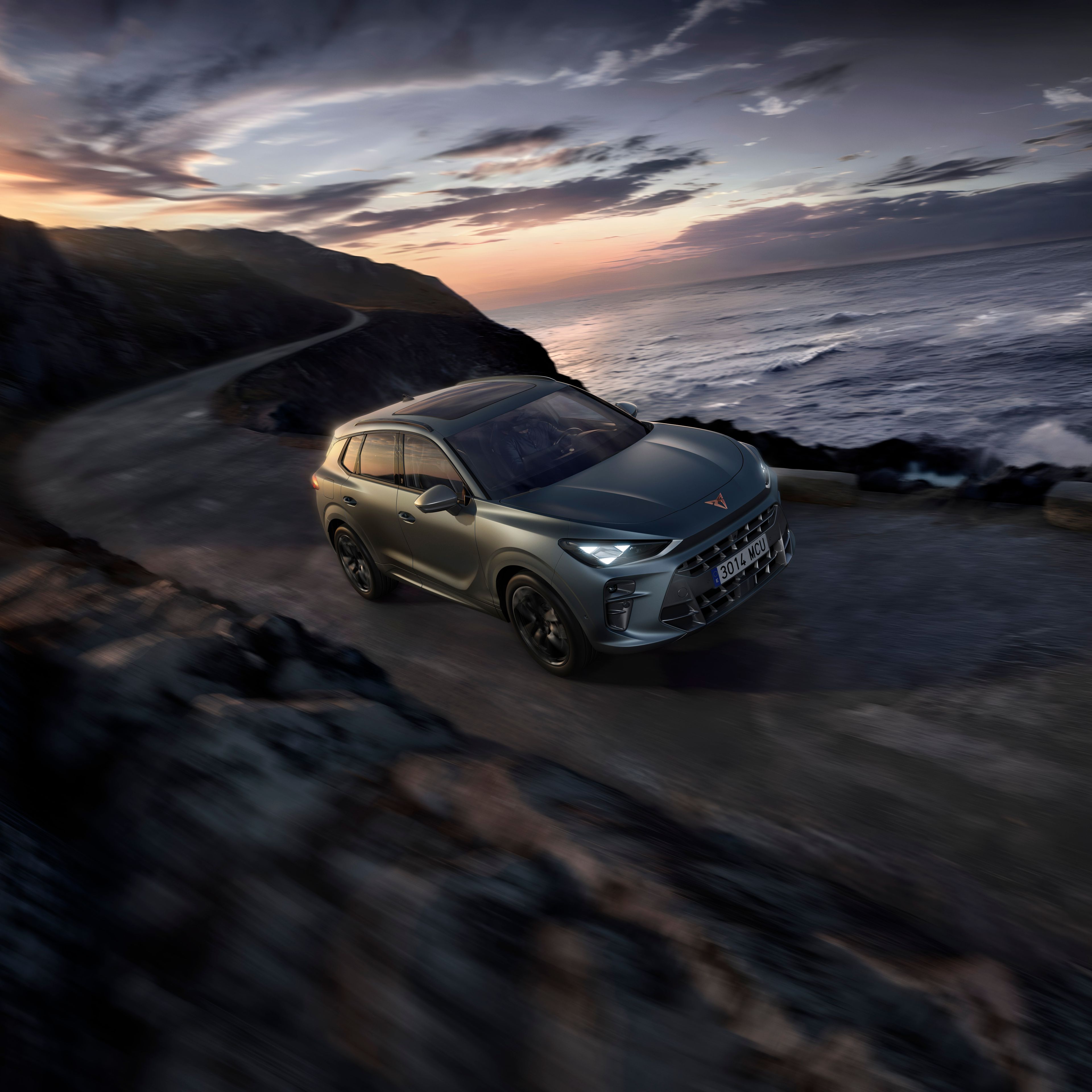 Automobil jazdí po úzkej ceste vedľa pobrežia za súmraku. More je rozbúrené a obloha je zamračená. Autu sa darí aj napriek náročným podmienkam na ceste.