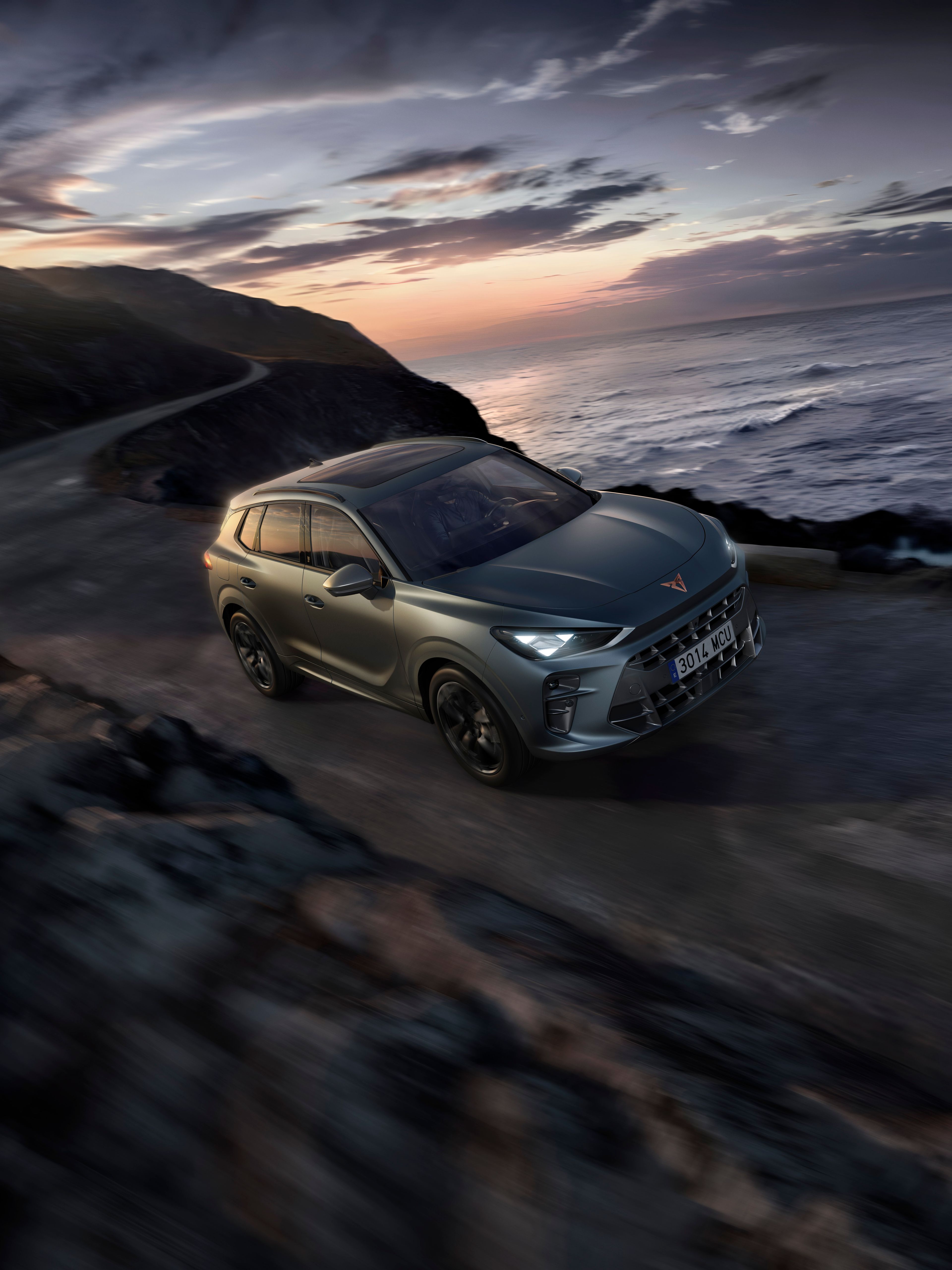 Automobil jazdí po úzkej ceste vedľa pobrežia za súmraku. More je rozbúrené a obloha je zamračená. Autu sa darí aj napriek náročným podmienkam na ceste.