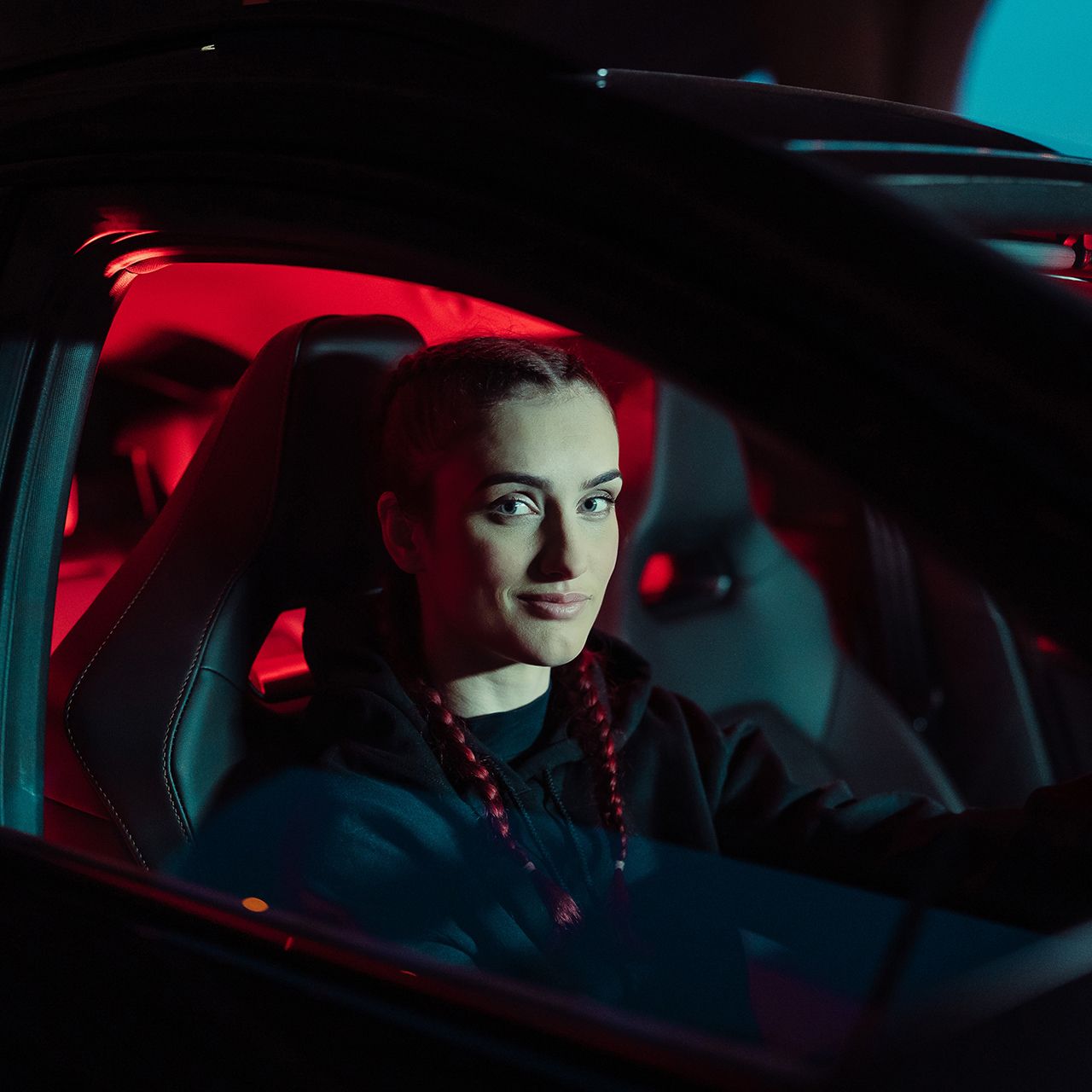 Žena sedí za volantom auta s červeným osvetlením interiéru. Má na sebe tmavé oblečenie a zapletené vrkoče. Pozadie je tmavé s modrým nádychom.