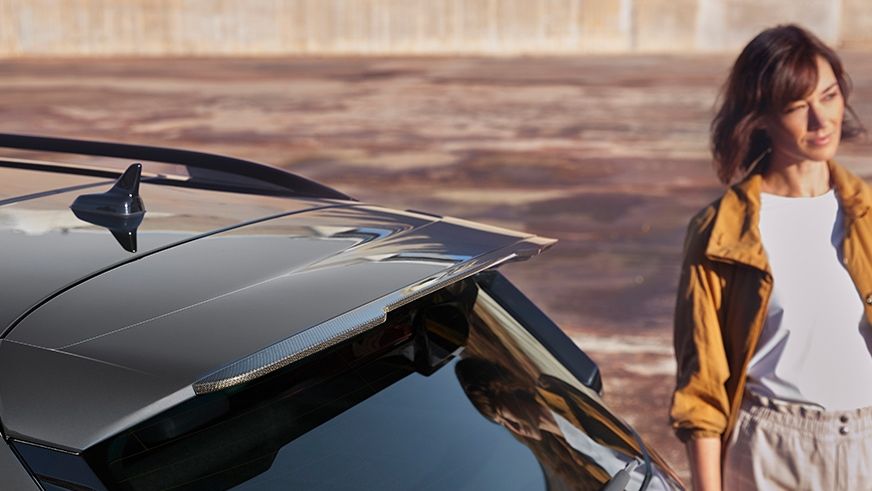 Žena stojí vedľa zaparkovaného auta s moderným dizajnom. Má na sebe biele tričko a žltú bundu. Pozadie je rozmazané.