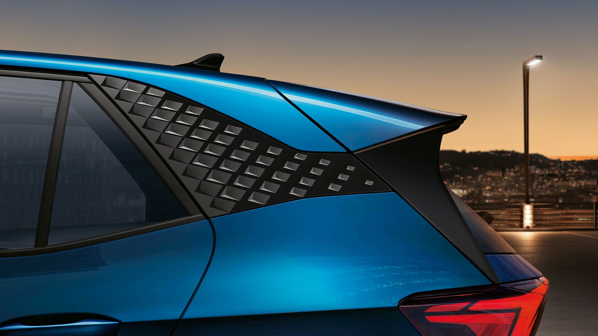 CUPRA Born v modrom lakovaní Aurora s dizajnovým zadným C stĺpikom. V pozadí je výhľad na nočné mesto pri západe slnka.