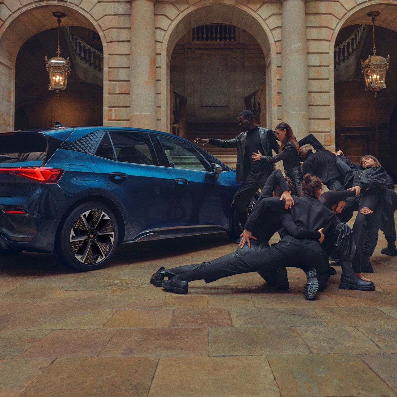 Modré auto je zaparkované na nádvorí historickej budovy. Skupina tanečníkov v čiernom oblečení vykonáva tanečné alebo dramatické vystúpenie okolo auta.