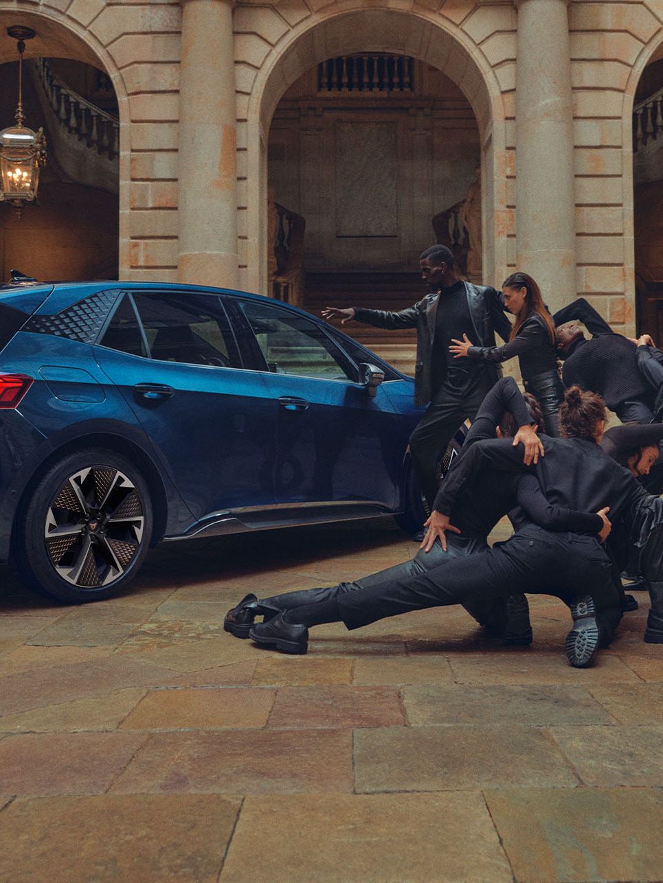 Modré auto je zaparkované na nádvorí historickej budovy. Skupina tanečníkov v čiernom oblečení vykonáva tanečné alebo dramatické vystúpenie okolo auta.