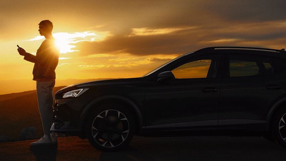 Osoba stojí vedľa SUV auta pri západe slnka. Pozadie je tmavé a obloha je zafarbená oranžovou farbou. Osoba má v ruke mobilný telefón.