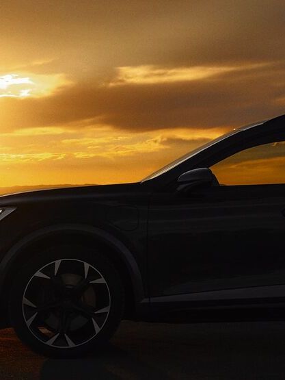 Osoba stojí vedľa SUV auta pri západe slnka. Pozadie je tmavé a obloha je zafarbená oranžovou farbou. Osoba má v ruke mobilný telefón.