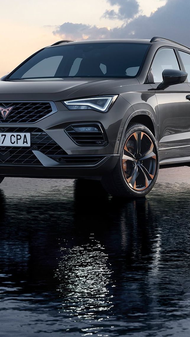 SUV značky Cupra stojí na povrchu vody pri západe slnka. Pozadie je zahalené do jemných odtieňov ružovej a modrej. Skvelý odraz vozidla na vode dodáva scéne pokojný a pôsobivý efekt.