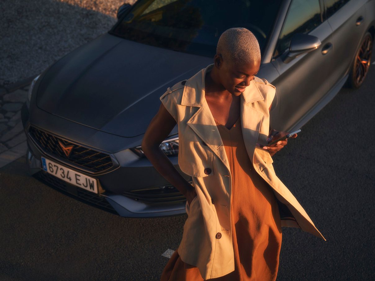 SUV stojí na parkovacom mieste na ceste. Od vozidla odchádza žena, ktorá drží mobilný telefón a pozerá sa na neho. Oblečené má oranžové šaty a béžovú vestu.
