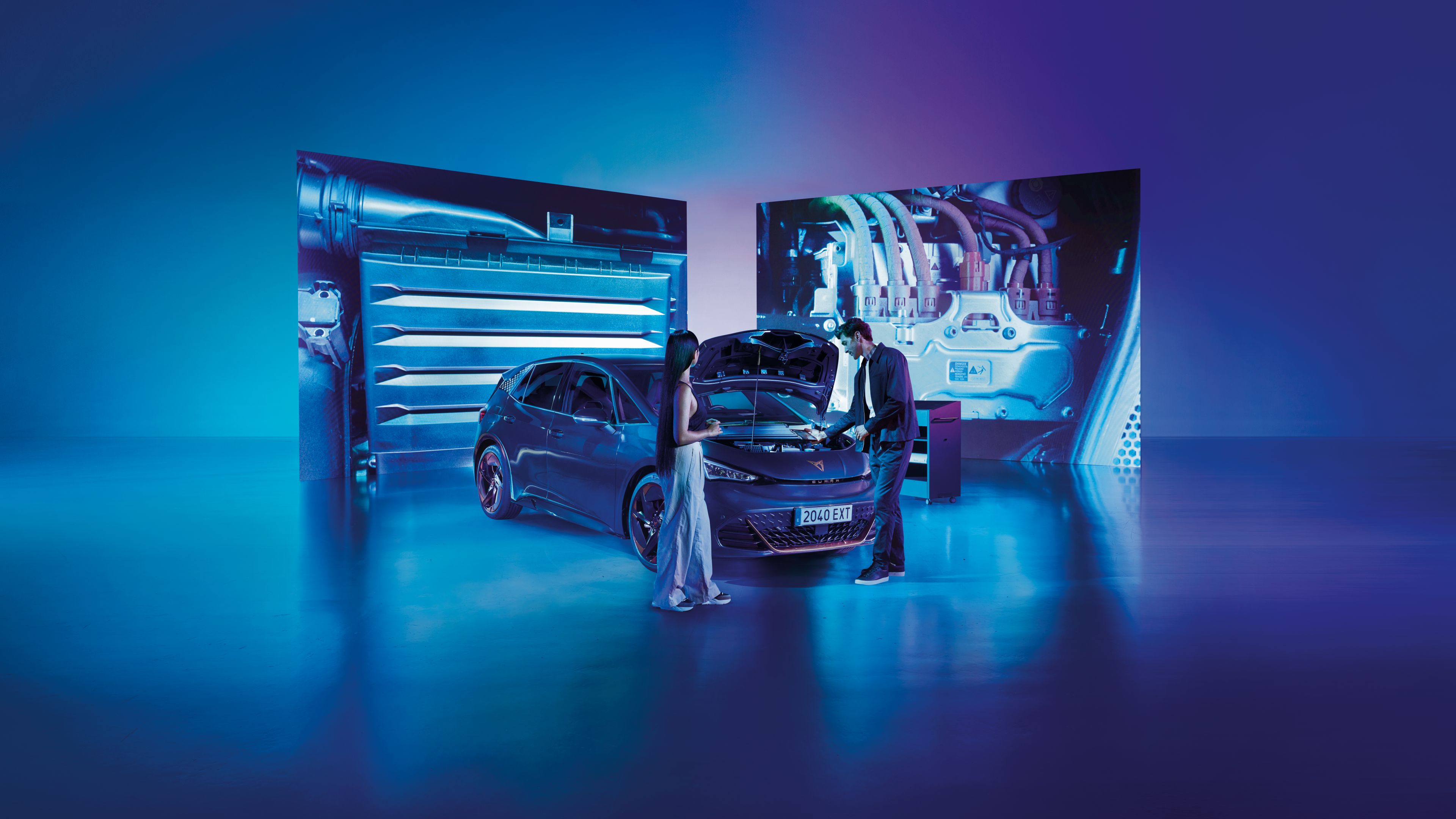 Muž stojí pri CUPRA Born s otvorenou kapotou a pozerá sa do priestoru motora. Žena stojí pri vozidle, za ktorým sú dve veľké obrazovky s detailom na rôzne časti motora. Pozadie tvorí modro-ružové osvetlenie.
