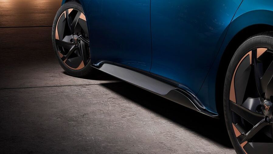 Detailný pohľad na bočnú časť modrého športového auta s elegantnými čiernymi a medenými diskami kolies. Vozidlo stojí na betónovom povrchu. Pozadie je tmavé s jemným osvetlením.