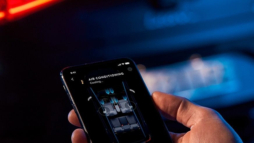 Ruka drží mobilný telefón, na ktorom je aplikácia na ovládanie klimatizácie v automobile. Na displeji telefónu je zobrazené rozhranie klimatizácie s možnosťou nastavenia teploty. V pozadí je rozostrený pohľad na zadnú časť automobilu.