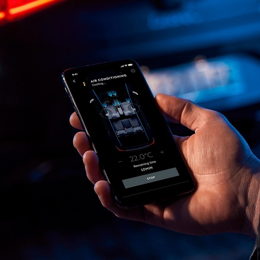 Ruka drží mobilný telefón, na ktorom je aplikácia na ovládanie klimatizácie v automobile. Na displeji telefónu je zobrazené rozhranie klimatizácie s možnosťou nastavenia teploty. V pozadí je rozostrený pohľad na zadnú časť automobilu.