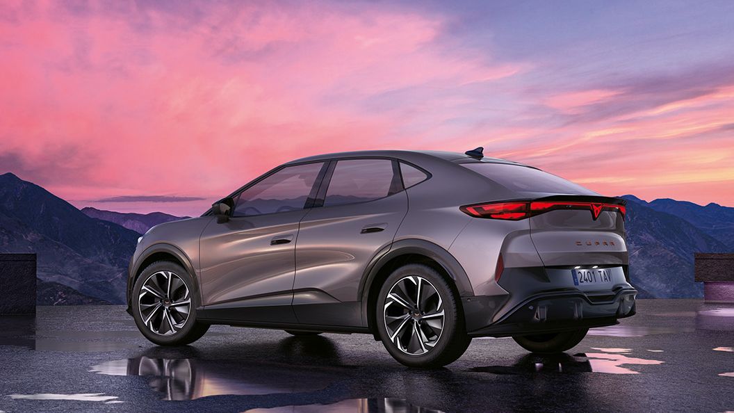 Moderné SUV so šedou metalízou je zaparkované na mokrej ploche s horami v pozadí. Obloha je sfarbená do ružova pri západe slnka. Auto má elegantný dizajn s aerodynamickými líniami.