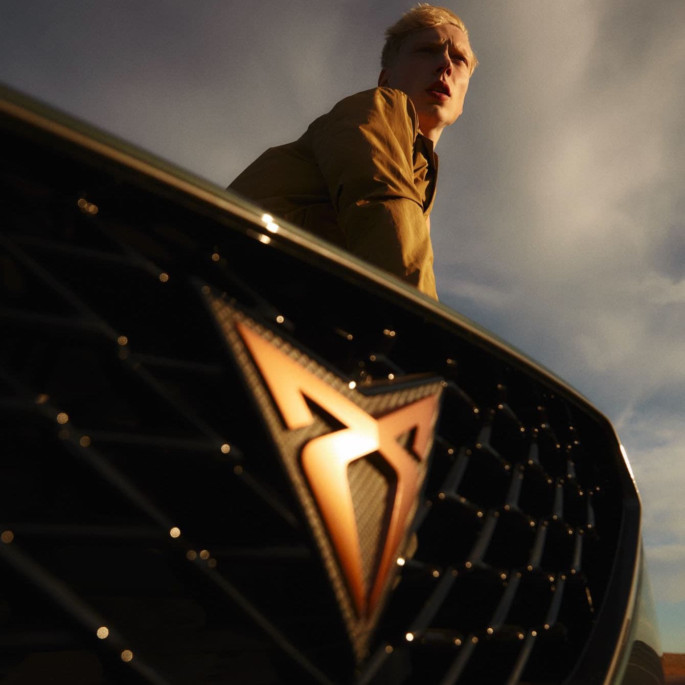 Detailný pohľad na prednú masku auta s logom CUPRA. V pozadí je postava muža.