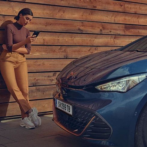 Žena stojí opretá o drevenú stenu a drží v ruke mobil. Vedľa nej je zaparkované športové auto.