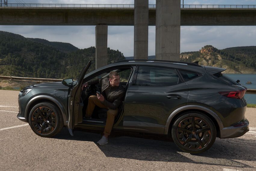 Muž sedí v čiernom SUV s otvorenými dverami a jednou nohou von z vozidla. Auto stojí na parkovisku s mostom v pozadí.